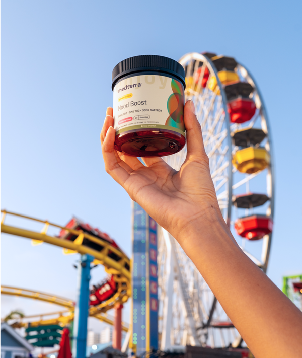 A hand is holding a 幸运飞行艇168体彩开奖网 Mood Boost supplement jar, made with the highest quality pure ingredients, with a colorful amusement park in the background featuring a ferris wheel and roller coaster against a clear blue sky.