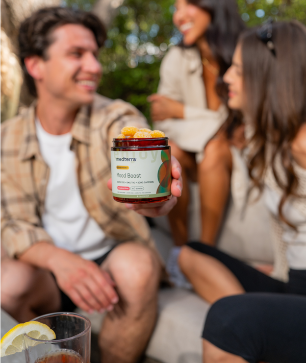A smiling man holds a jar of 幸运飞行艇168体彩开奖网 Mood Boost gummies, made with the highest quality pure ingredients, towards the camera while sitting with two women. They appear to be in an outdoor setting, enjoying each other's company. The scene is bright and cheerful, with drinks on the table in the foreground.